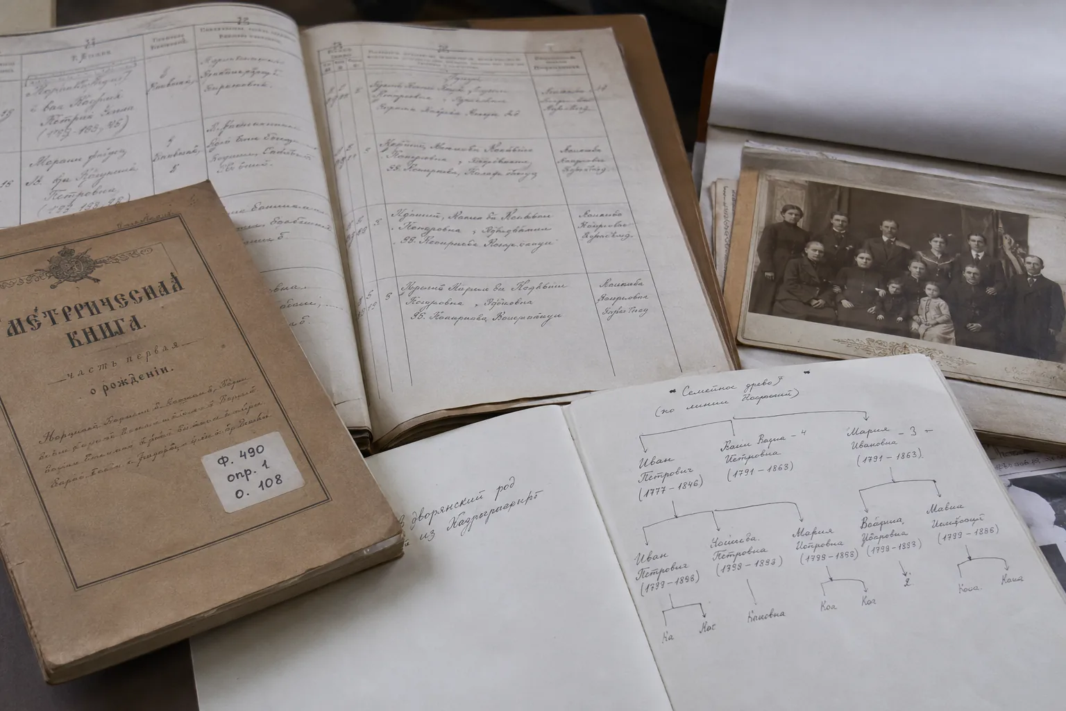 Open archival record books, a handwritten family tree chart, and an old family photograph laid out on a desk